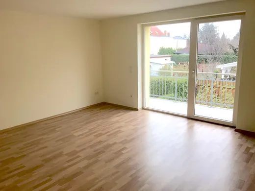 Wohnzimmer mit breiter Balkontür, dahinter ein Balkon mit Blick auf eine Hecke und die Nachbarhäuser.