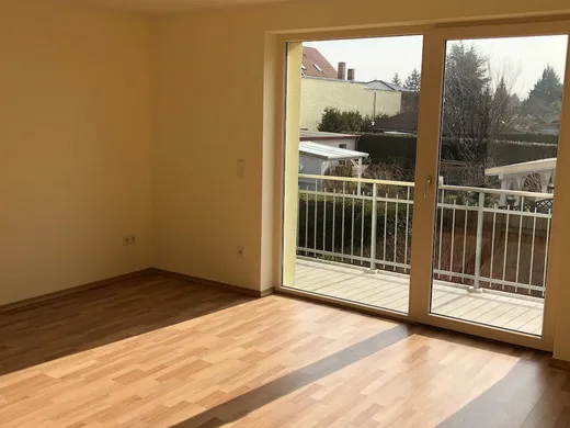 Leeres Wohnzimmer mit Laminatboden, warm gestrichenen Wänden und einer bodentiefen Balkontür, durch die Sonne hereinfällt.
